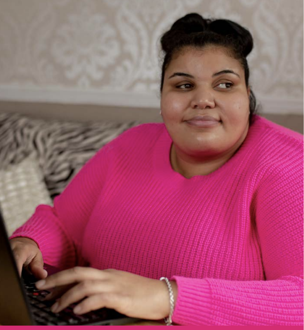 Woman with dark hair tied up on top of head. She is wearing a pink jumper and using a laptop.